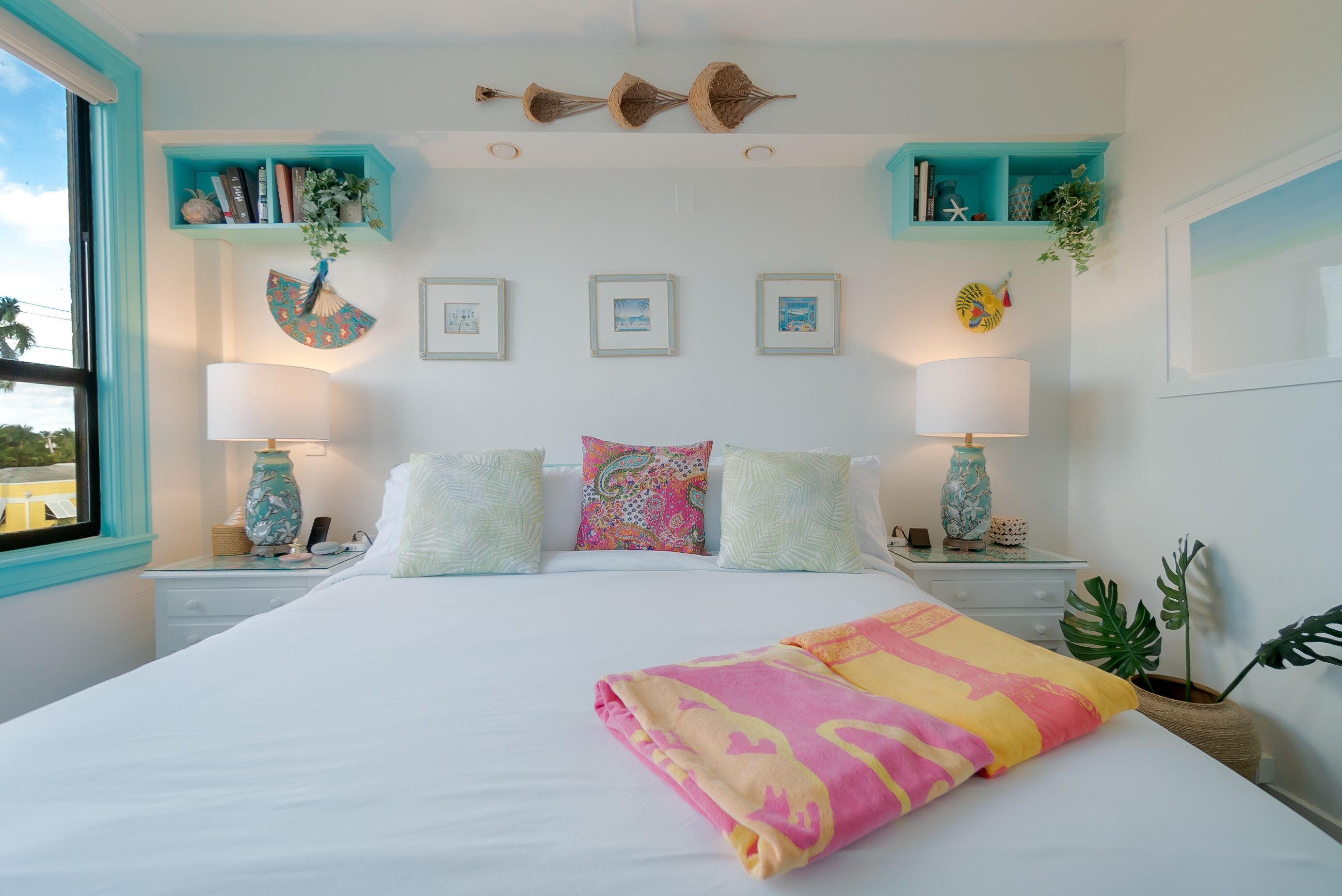 Una cama bien hecha con una manta rosa y amarilla, dos mesitas de noche con lámparas, y estantes en la pared con libros y plantas.