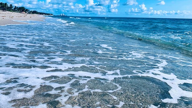 Una playa con agua cristalina, rocas a la vista y una costa lejana con edificios.
