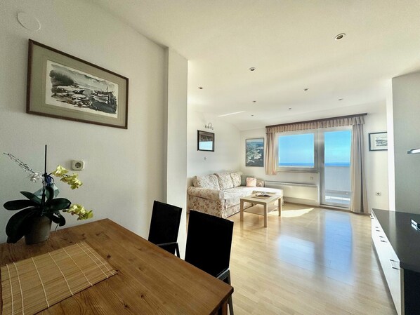 A modern living room with a dining area, a sofa, a coffee table, and a view of the sea through a large window.