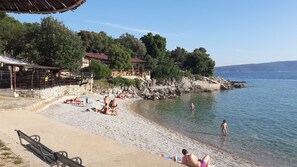 Ein Kiesstrand mit badenden und sonnenden Menschen, eine hölzerne Konstruktion mit rotem Dach und eine felsige Küste mit Bäumen.