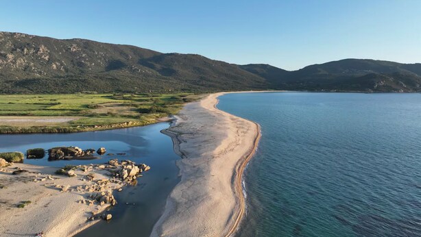 Paesaggio costiero con spiaggia sabbiosa, mare calmo e colline verdi sullo sfondo.