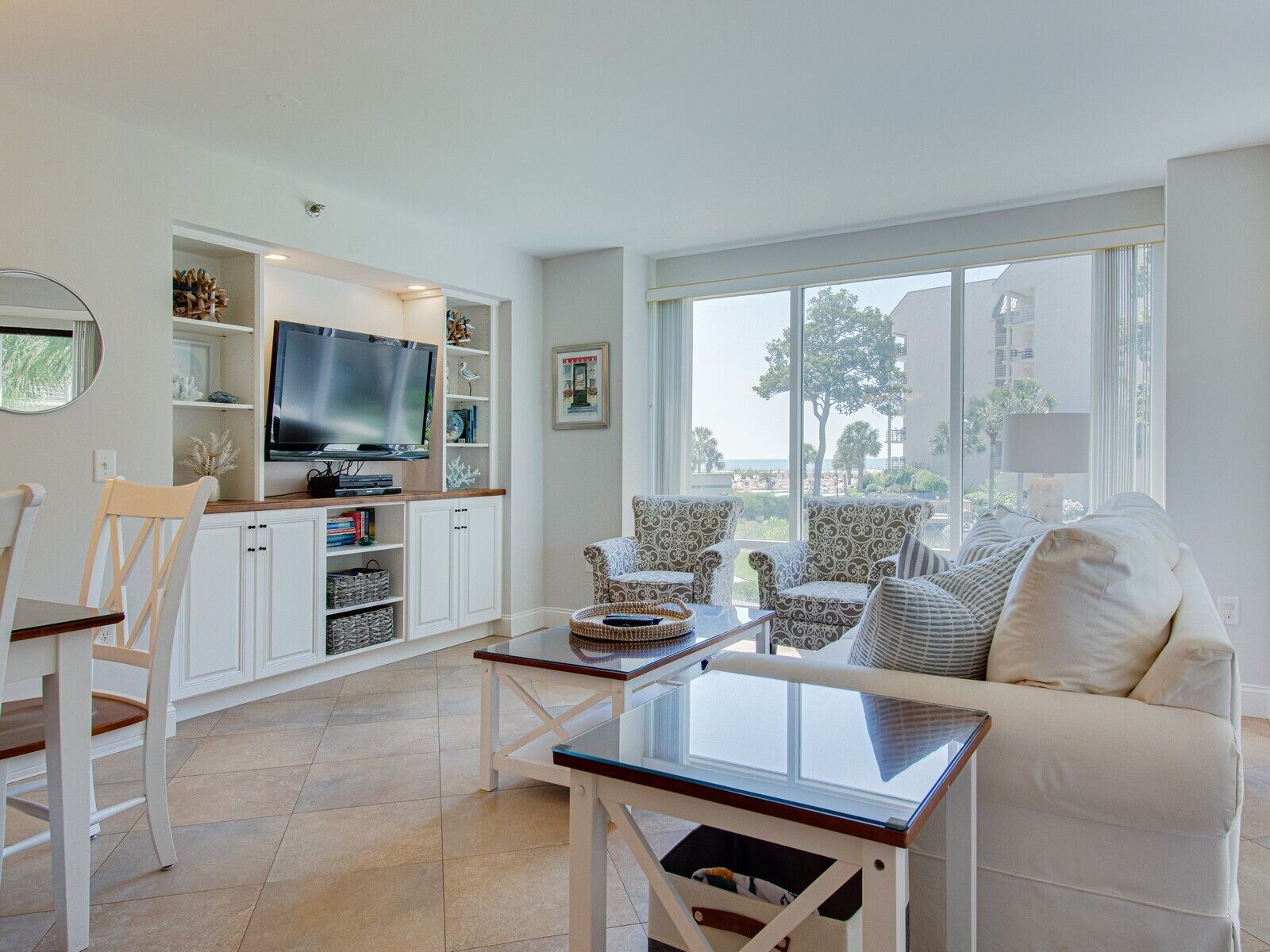 A modern living room with a TV, white furniture, and large windows offering a view of the outdoors.