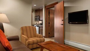 A living room with a sofa, armchair, and coffee table. A TV is mounted on the wall, and a kitchen is visible through an open door.
