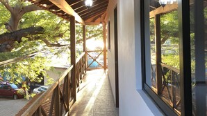 A covered corridor with wooden beams, a tiled floor, and large windows offering views of trees and parked cars.