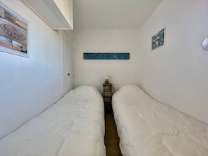 Un pasillo estrecho con dos camas individuales, una mesita con lámpara y un cartel que dice 'VACANCES'.