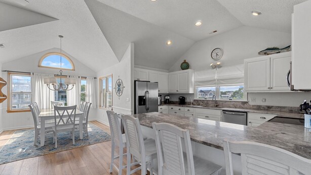 A spacious kitchen with white cabinetry, a large island, and a dining area with white chairs. - 1522 - Sand Dollar - Oceanside Corolla Home w/ Pool & Volleyball (Corolla)