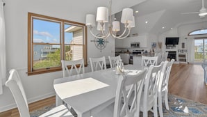 A spacious dining area with a large table and white chairs, a chandelier, and a view of the outside. - 1522 - Sand Dollar - Oceanside Corolla Home w/ Pool & Volleyball (Corolla)