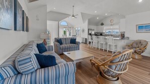 A spacious living room with striped sofas, a coffee table, and wicker chairs. A kitchen and dining area are visible in the background. - 1522 - Sand Dollar - Oceanside Corolla Home w/ Pool & Volleyball (Corolla)