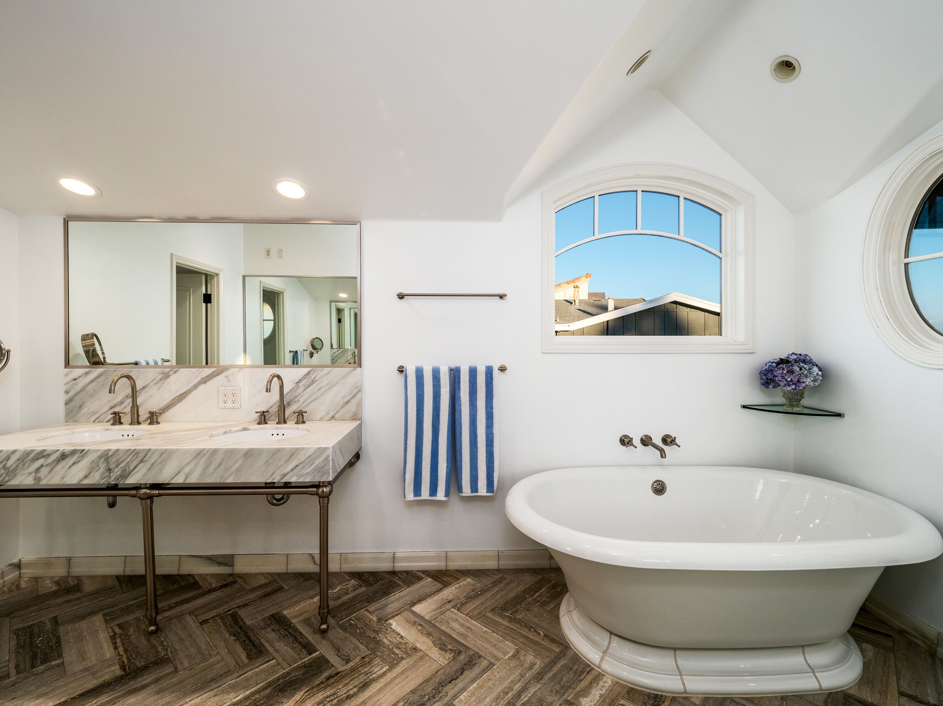 Un baño con lavamanos de mármol, una bañera independiente, un espejo grande y una ventana con vistas a edificios.
