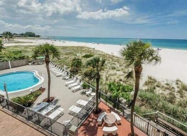 Zona de piscina con sillas de descanso y una mesa, con vista a una playa con palmeras.