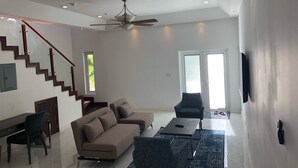 A spacious living room with a high ceiling, a ceiling fan, a staircase with wooden railings, a sofa, two armchairs, a coffee table, and a TV mounted on the wall.
