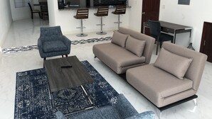 A modern living room with a sofa, armchair, and coffee table. There are two bar stools and a dining table with chairs in the background.