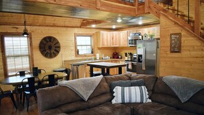 A cozy cabin interior with a brown leather couch, wooden dining table, and a kitchen area with wooden cabinets and a stainless steel refrigerator. - Majestic Moose Lodge- Oark Cabin Rentals (Oark)