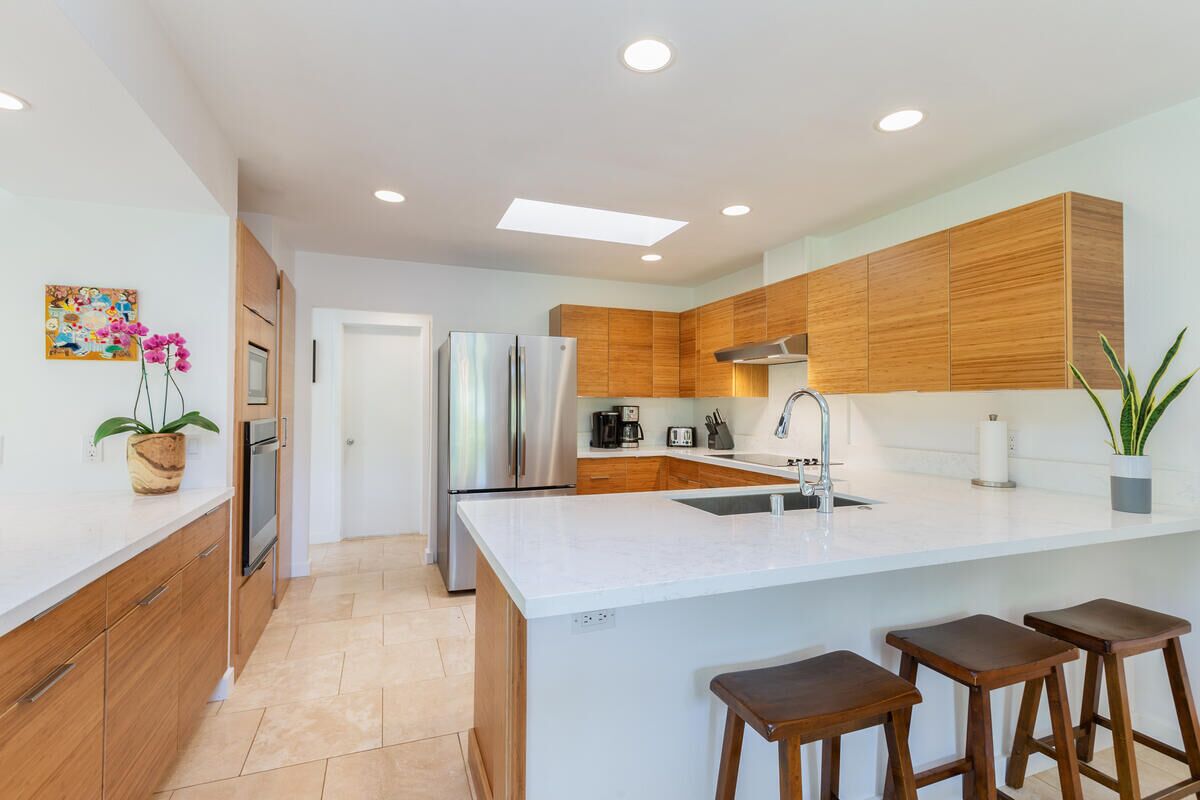 A modern kitchen with wooden cabinets, a stainless steel refrigerator, a white countertop, and a bar stool area.