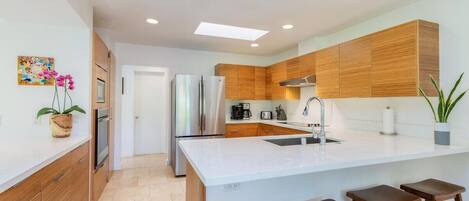 A modern kitchen with wooden cabinets, a stainless steel refrigerator, a white countertop, and a bar stool area.