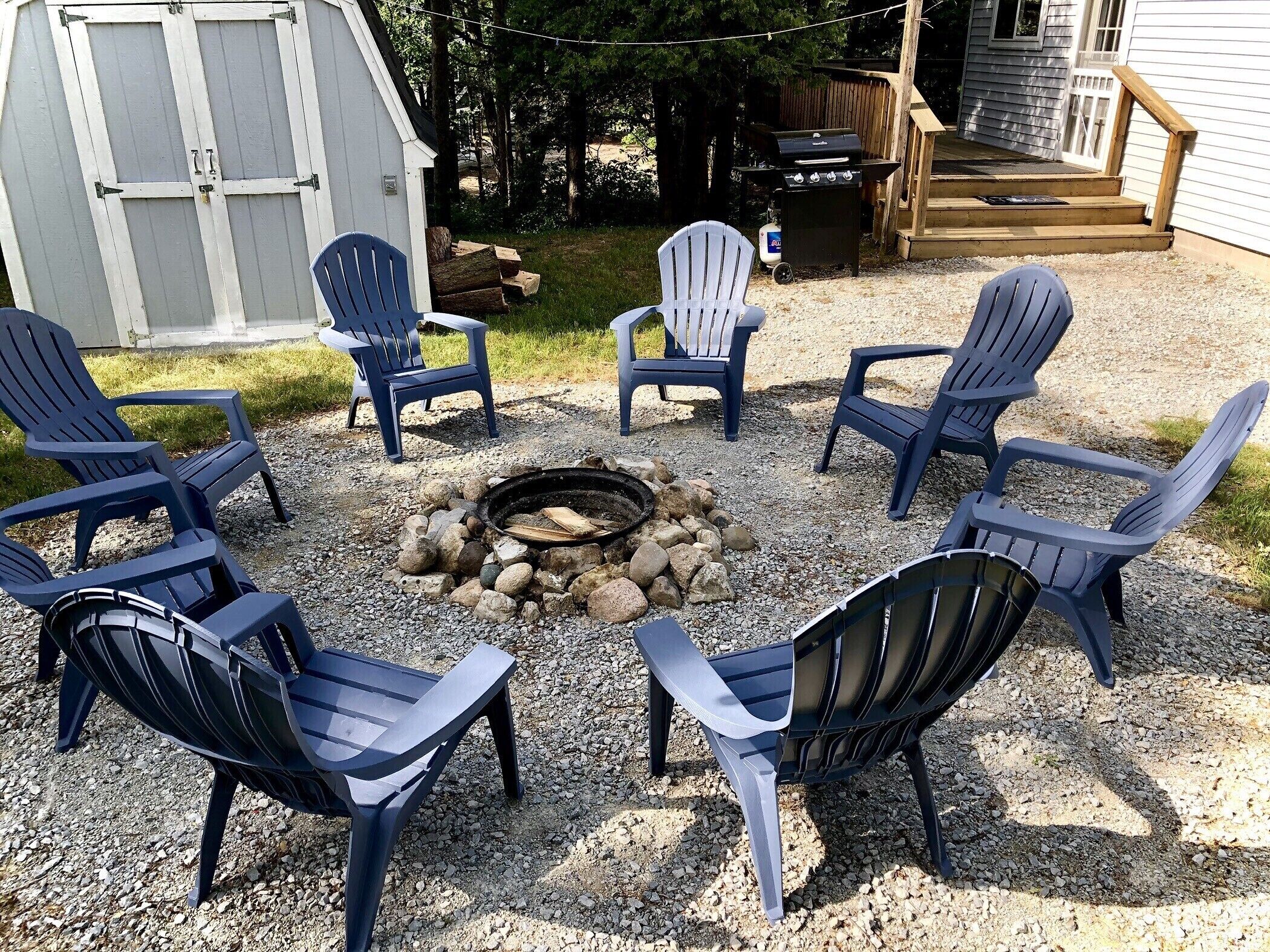 A fire pit surrounded by six Adirondack chairs in a gravel area.