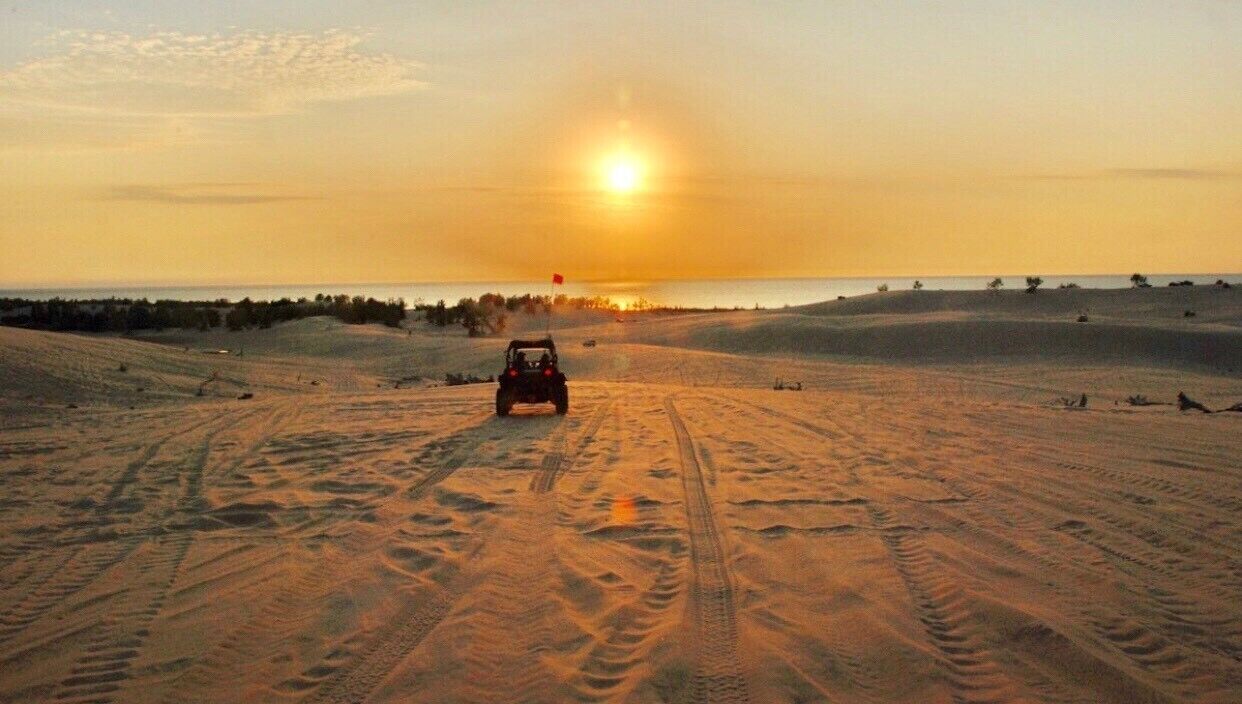 A desert landscape with a vehicle driving on sand dunes during sunset.