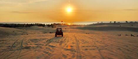A desert landscape with a vehicle driving on sand dunes during sunset.