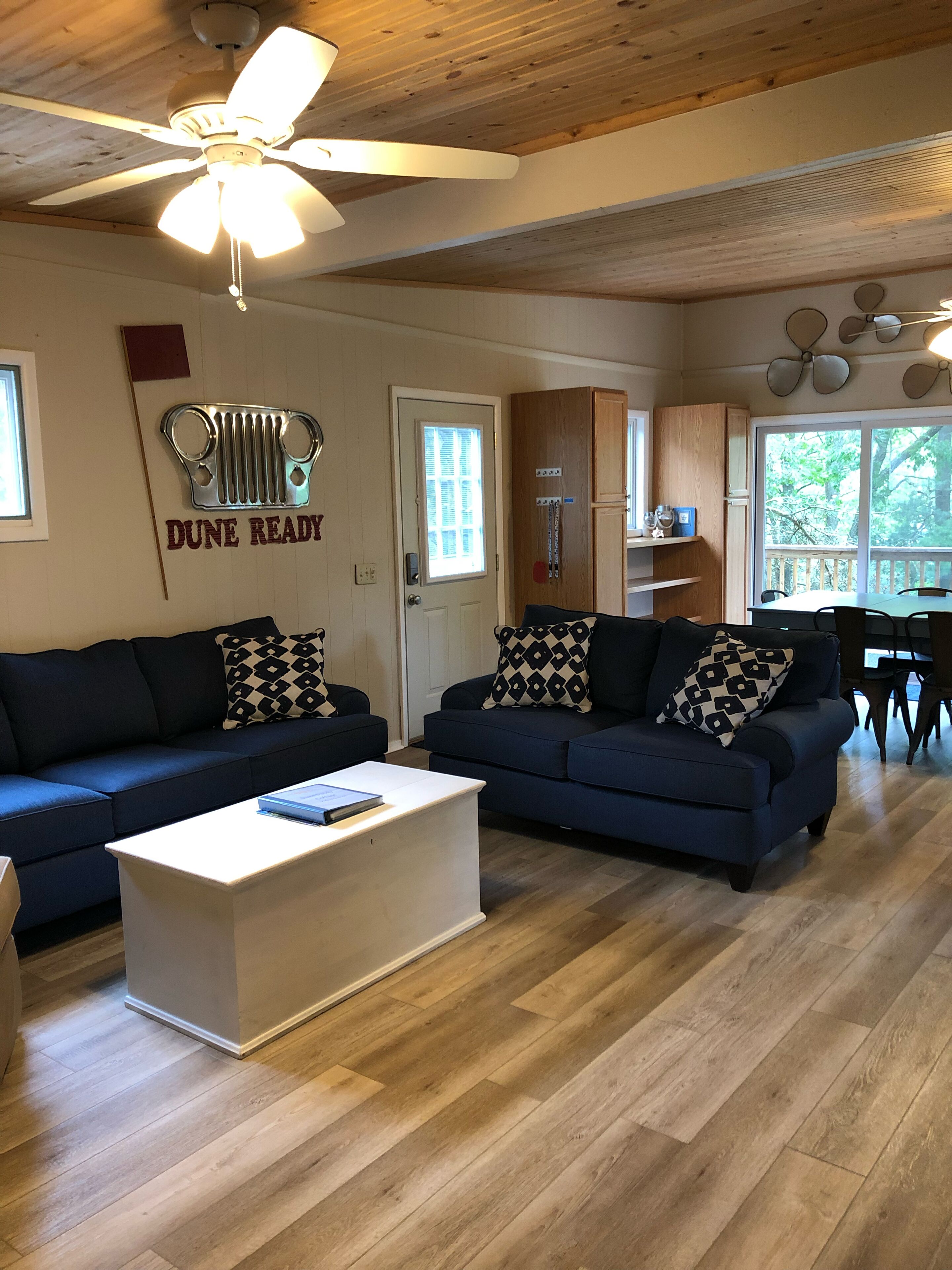 A living room with two dark blue sofas, a white coffee table, and a ceiling fan. There is a wooden ceiling and a window with a view of trees.