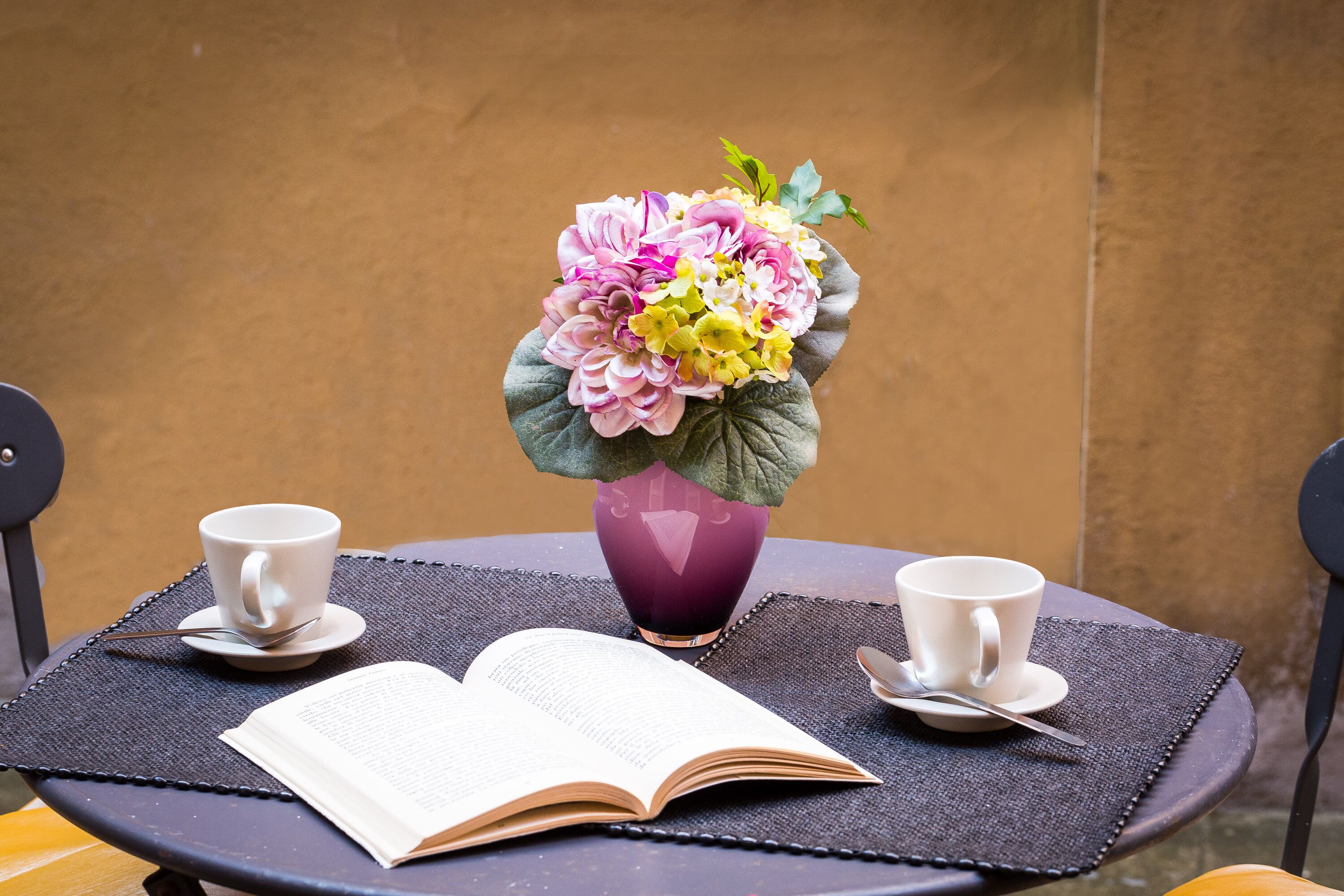 Une table ronde sur laquelle se trouve un vase violet contenant un bouquet de fleurs, deux tasses blanches et un livre ouvert.