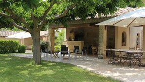 An outdoor patio area with a stone table and chairs, a pergola providing shade, and a view of a building with a tiled roof. - 360 m2 house (Meynes)