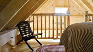 Un dormitorio acogedor en el ático, con techo inclinado, un banco de madera, una cama con colcha a rayas y vistas al exterior a través de una ventana.