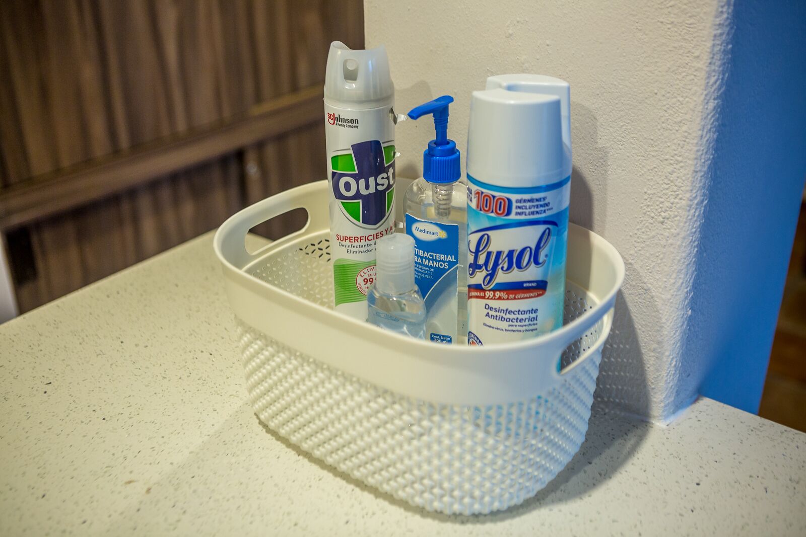 Una cesta blanca contiene una botella de Oust, una botella de Lysol y una botella de Medimart sobre una encimera con motas.