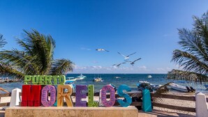 Een bord aan het strand met kleurrijke letters waarop 'Punto Morelos' staat, met op de achtergrond de zee, boten en palmbomen.