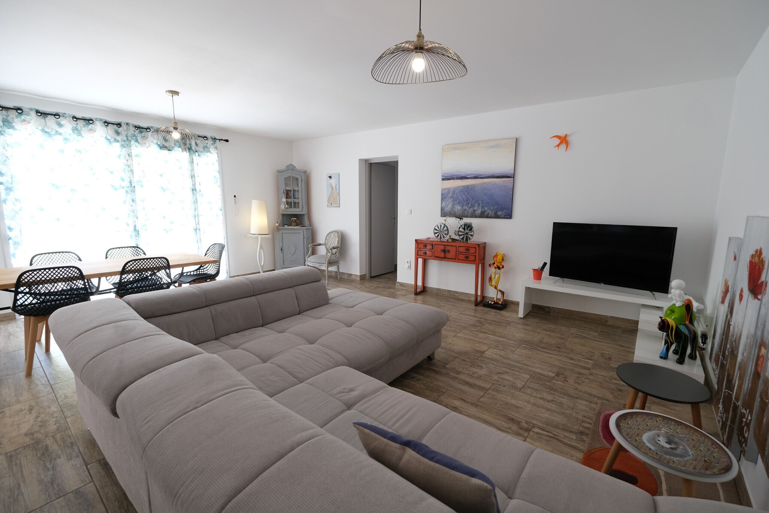 A modern living room with a large sectional sofa, a dining area with chairs, a TV, and artwork on the walls.