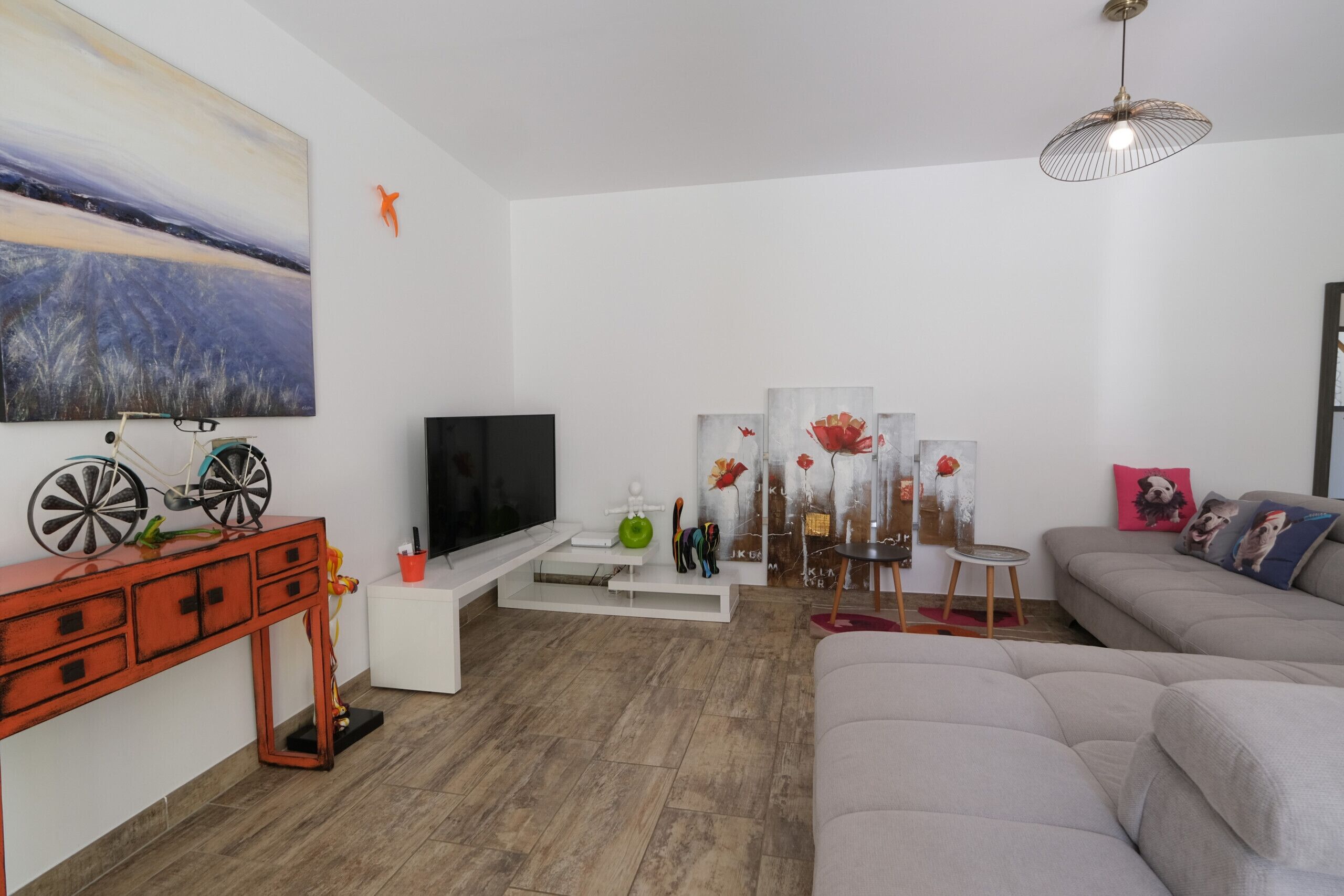 A modern living room with a flat-screen TV, a red console table, a grey sofa, and a painting of a landscape.