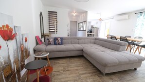 A modern living room with a large L-shaped sectional sofa, a small round table, and a coffee table. There is a dining area with a wooden table and chairs. The room has a neutral color palette and features artwork on the walls.
