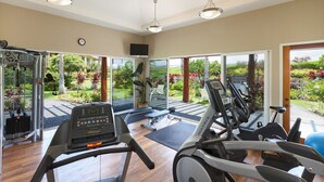 Ein Fitnessraum mit Laufbändern, einem Ergometer und einem Kraftgerät. Große Fenster bieten Blick auf einen Garten.