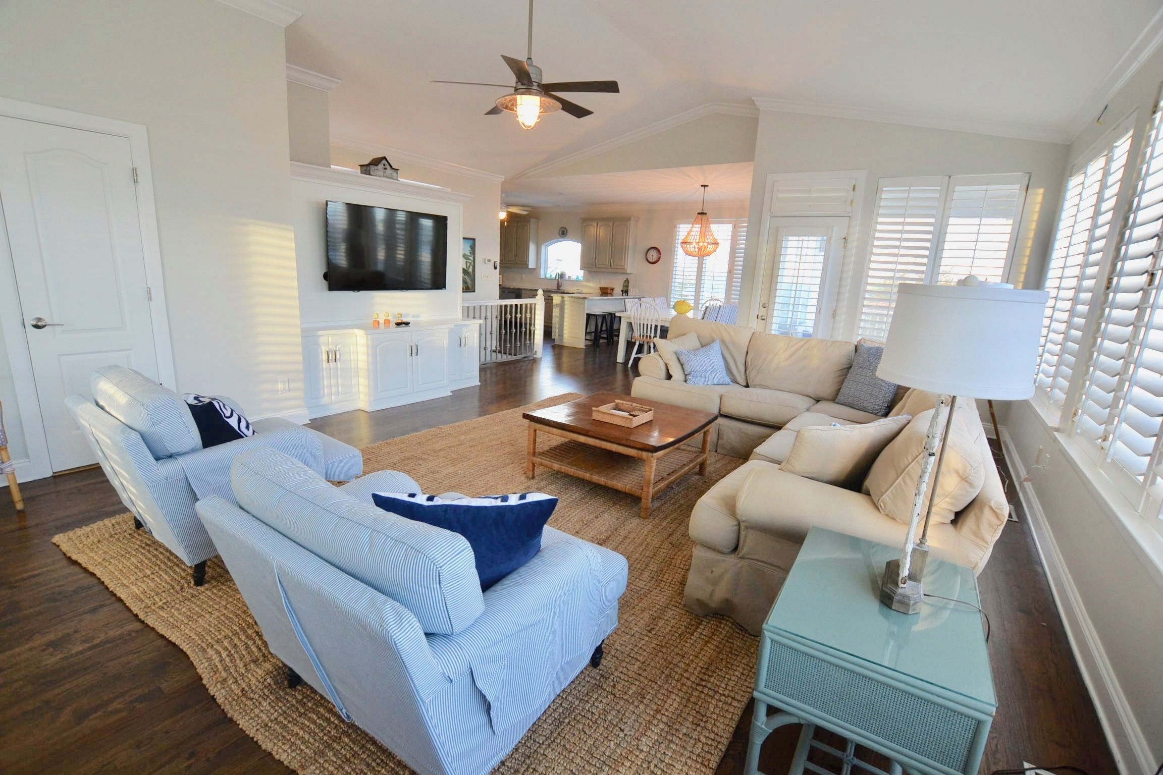 A spacious living room with a sofa, armchairs, and a coffee table. A television is mounted on the wall, and there is a ceiling fan.