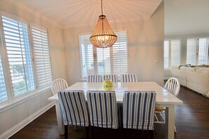 Un coin repas avec une table blanche et des chaises à rayures, un luminaire suspendu et de grandes fenêtres équipées de stores.