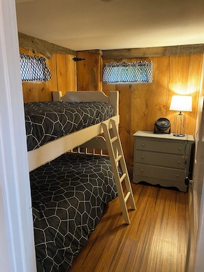 Un lit superposé avec une échelle, une table de chevet avec une lampe et une commode dans une pièce aux murs et au plancher en bois.