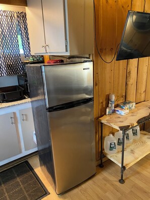 A stainless steel refrigerator with a freezer compartment, a wooden table with a television, and a kitchen area with cabinets and a patterned curtain.