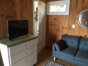 Quarto com televisão sobre bancada, sofá cinza, paredes de madeira, uma porta e uma janela.