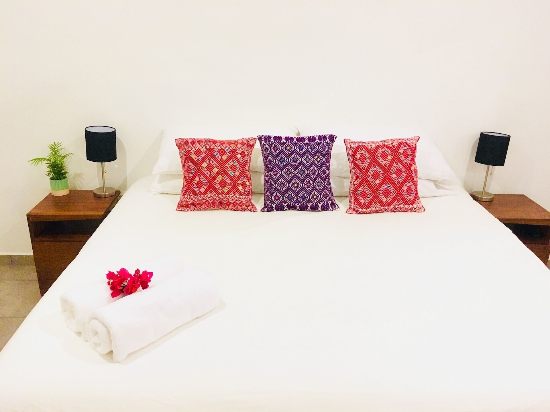 Una cama con tres almohadones estampados, dos mesitas de noche con lámparas, una planta pequeña y una toalla blanca enrollada con flores rojas.