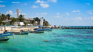 A coastal scene with boats docked at a pier, a lighthouse, and a beach with palm trees. - AQUA GUESTHOUSE 3~ STEPS FROM THE BEACH ~ POOL ~ A+ INTERNET (Puerto Morelos,)