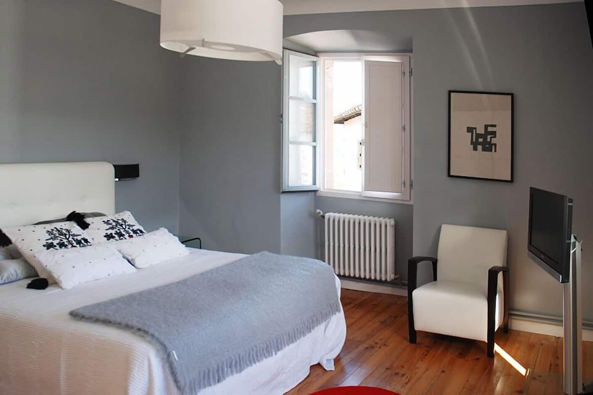 A bedroom with a bed, a chair, a radiator, a window, and a TV.
