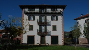 A two-story building with a balcony on the upper floor, a central entrance with double doors, and a small garden area in front. - Zilbetinea (Baztan)