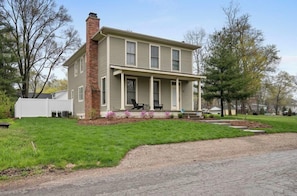 A two-story house with a brick chimney, a covered porch with chairs, and a well-maintained lawn.