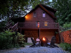Una cabaña de madera con base de piedra, un balcón y una zona con fogón y dos sillas Adirondack.