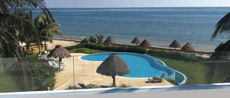 A pool area with a clear view of the ocean, surrounded by palm trees and thatched umbrellas.