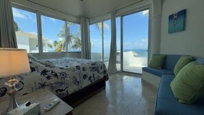Habitación de hotel con cama, escritorio con lámpara, control remoto y sofá azul. La habitación tiene ventanas amplias con vista a la playa y palmeras.