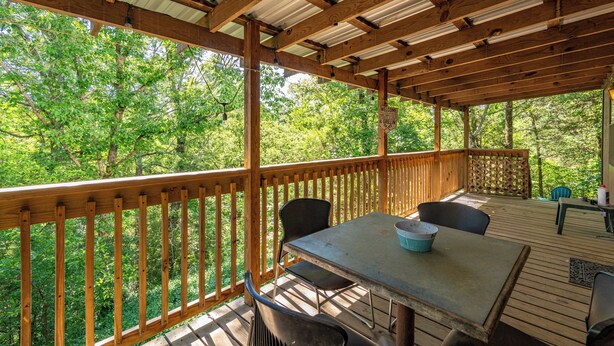 A wooden deck with a table and chairs, surrounded by a wooden railing, overlooking a forested area. - The Enchanted — A Quiet Riverside Retreat in the Heart of the Ouachita (Pencil Bluff)