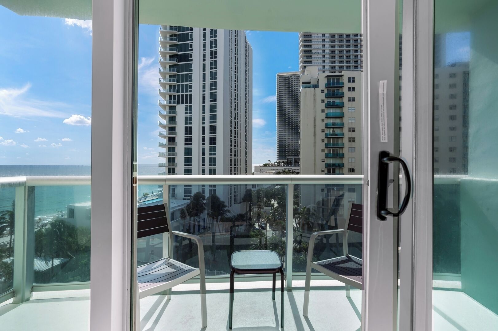 A balcony with a glass door, two chairs, and a view of tall buildings and a body of water.