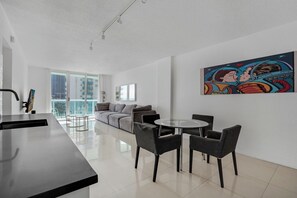 Una sala moderna con un sofá, una mesa de comedor con sillas, una zona de cocina con un fregadero y un ventanal que ofrece vistas a edificios.