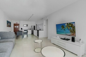 A modern living room with a flat-screen TV, a sofa, a dining area, and a kitchen.