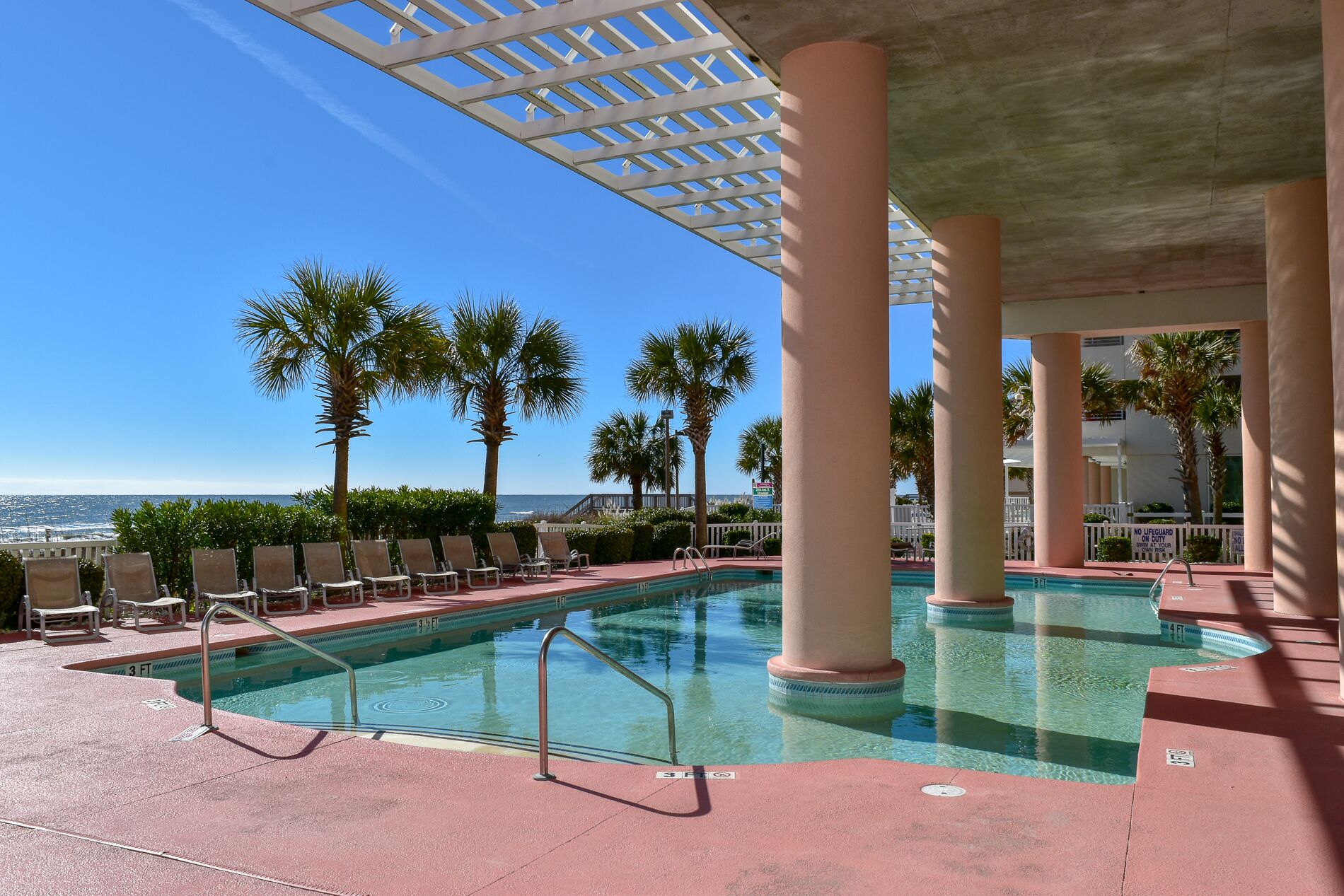 A pool area with lounge chairs, palm trees, and a view of the ocean.